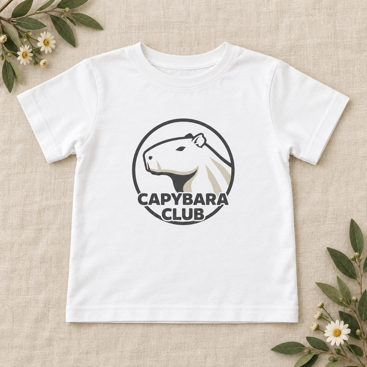 AI-created flat lay: White Capybara Club toddler tee with dark artwork on cream linen and green foliage.