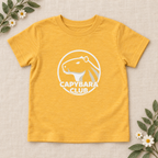 AI-created flat lay: Heather Yellow Gold Capybara Club toddler tee with white artwork and botanical styling.