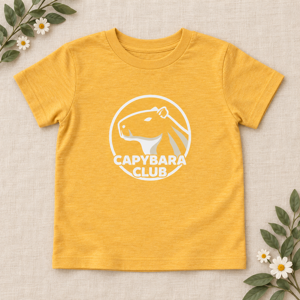 AI-created flat lay: Heather Yellow Gold Capybara Club toddler tee with white artwork and botanical styling.