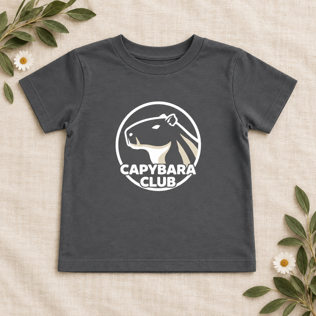 AI-created flat lay: Asphalt Capybara Club toddler tee with white artwork and botanical styling.
