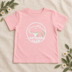 AI-created flat lay: Pink Capybara Club toddler tee with white artwork and botanical styling.