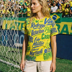 Woman wearing a yellow sports jersey with text in front of a soccer goal and crowd.