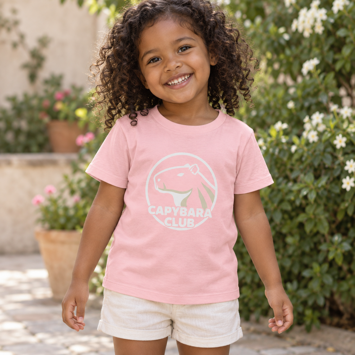 AI-created lifestyle mockup: Pink Capybara Club toddler tee in a garden courtyard.