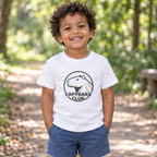 AI-created lifestyle mockup: White Capybara Club toddler tee on a child in a leafy park.